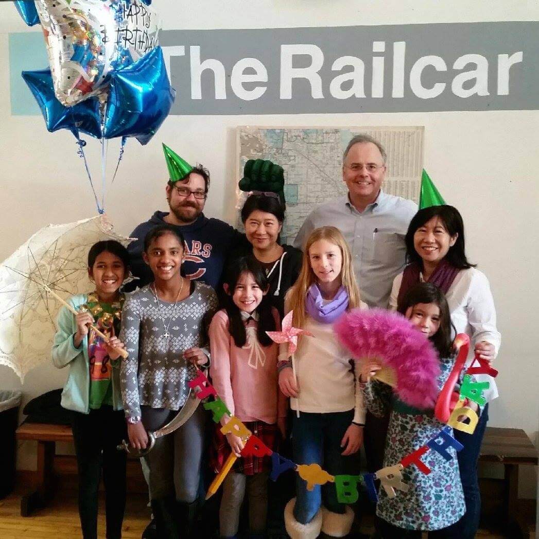 Escape room team adorning festive birthday accessories poses in front of The Railcar logo