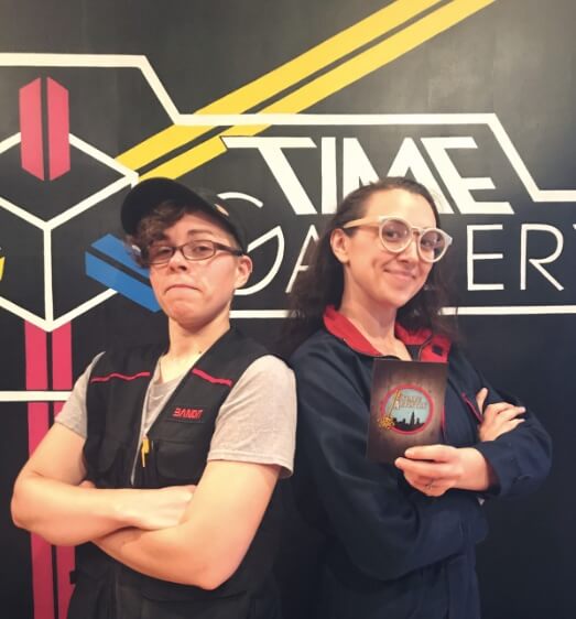 Escape Artistry Owner And Game Designer Pose In Front Of Time Gallery Logo